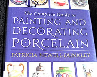 Op de afbeelding: Een boek getiteld "The Complete Guide to Painting and Decorating Porcelain" door Patricia Newell-Dunkley. De blauwe omslag met witte tekst toont afbeeldingen van gedecoreerde porseleinen borden en vazen.
