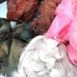 May include: A close-up of various colorful wool roving fibers. The colors include shades of pink, brown, green, and gray. Some fibers have metallic strands mixed in, adding a touch of sparkle. Ideal for fiber art projects.