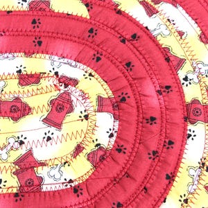 May include: Circular pet mat with a red, yellow, and white design. The mat features red fire hydrants, paw prints, and bone patterns. The mat is made of fabric and has a stitched construction.
