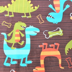 May include: Brown fabric with a repeating pattern of colorful cartoon dinosaurs and bone shapes. The dinosaurs are in shades of green, blue, and orange, with striped details. The bones are white and yellow.