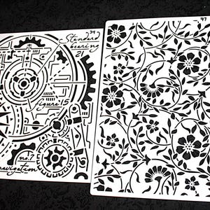 May include: Two white stencils with black designs. One features gears, a compass, and text including "navigation" and "figure 15." The other has a floral pattern with flowers and leaves. Both are on a black surface.