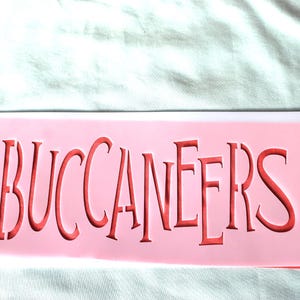 Op de afbeelding: Een roze sjabloon met het woord "BUCCANEERS" uitgesneden in wit.