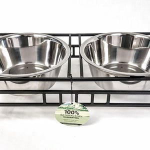 May include: A black metal double dog bowl stand with two stainless steel bowls. The bowls are round and sit in a rectangular frame. A tag with text hangs from the stand.