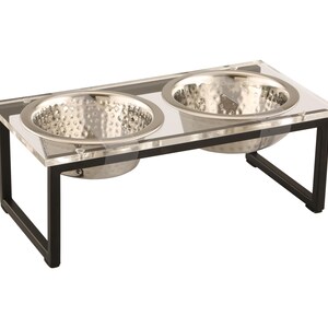 Puede incluir: Un soporte para cuencos de comida y agua para mascotas de acr&iacute;lico transparente con dos cuencos de acero inoxidable. El soporte tiene un marco de metal negro.