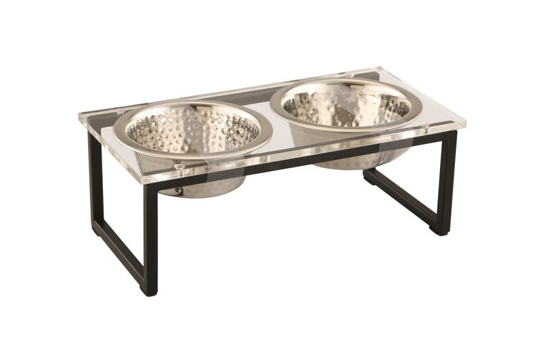 Puede incluir: Un soporte para cuencos de comida y agua para mascotas de acr&iacute;lico transparente con dos cuencos de acero inoxidable. El soporte tiene un marco de metal negro.