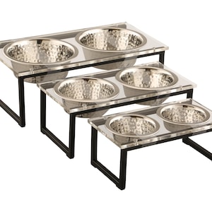 Puede incluir: Tres cuencos elevados para comida y agua para mascotas. Los cuencos est&aacute;n hechos de acero inoxidable y est&aacute;n colocados en plataformas de acr&iacute;lico transparente con marcos de metal negro.