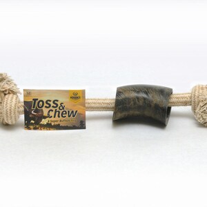 May include: A natural rope dog toy with a brown and green buffalo horn in the center. The rope is knotted at both ends and the toy is labeled "Toss & Chew - A Super Buffalo Toy".
