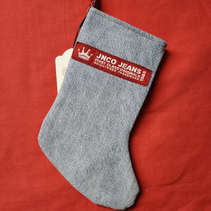 May include: A blue denim Christmas stocking with a red patch that says "JNCO JEANS FIRST CLASS ORIGINALS 1985 REGISTERED TRADEMARK" and a crown logo.