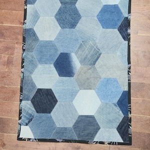 May include: A patchwork quilt featuring a pattern of blue denim hexagons. The quilt has a black border with a floral design. The denim shades range from light to dark blue, creating a textured, geometric design.