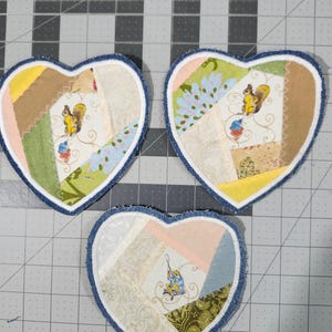 May include: Three heart-shaped fabric coasters. Each coaster features a patchwork design with various colors and patterns, including floral and squirrel motifs. The edges are trimmed with a blue denim fabric.