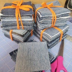 May include: Two stacks of denim fabric squares tied together with orange yarn. A single square of denim fabric is lying on a blue cutting mat next to a pair of pink scissors.