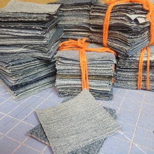 May include: A stack of denim fabric squares tied with orange string. The squares are arranged in a pyramid shape. There are several stacks of squares, and one stack is laying on its side. The squares are all the same size and are made of a light blue denim fabric.