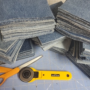 May include: Stacks of square denim fabric pieces in various piles. A yellow rotary cutter and orange scissors are on a blue cutting mat. The rotary cutter has the text "OLFA ROTARY CUTTER".