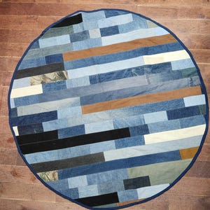 May include: Circular denim patchwork rug with a dark blue border. The rug is made from various shades of blue denim, with brown and white accents. The design is a mix of light and dark denim pieces.