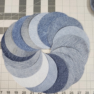May include: Circular arrangement of denim fabric pieces in varying shades of blue and gray. The pieces are arranged in a spiral pattern, creating a visually appealing design. The image is taken on a cutting mat with measurement markings.