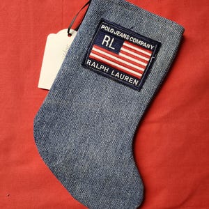 May include: Denim Christmas stocking with a patch featuring the text "POLO JEANS COMPANY" and "RALPH LAUREN" with a red, white, and blue flag design. The stocking has a red loop for hanging and a white tag.