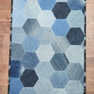 May include: A rectangular quilt composed of denim hexagons in various shades of blue. The quilt is bordered with a black fabric featuring a floral pattern. The patchwork design creates a visually appealing texture.