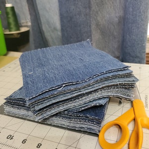 May include: A stack of denim fabric squares, cut and ready for sewing, sits on a gray cutting mat with a ruler and a pair of orange Fiskars scissors.