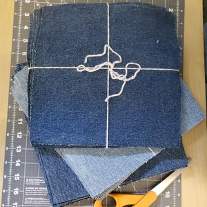 May include: A stack of blue denim fabric squares tied together with white string, and a pair of orange-handled scissors on a black cutting mat.
