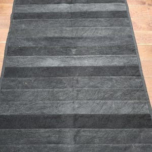 May include: A rectangular bath mat featuring a horizontal striped pattern in alternating shades of charcoal gray and black. The mat has a soft, textured surface and is finished with a solid black fabric border along all four edges.