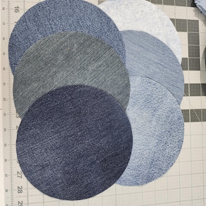 May include: A collection of denim fabric circles in various shades of blue, arranged on a gray cutting mat. The circles range from light to dark denim, showcasing different washes and textures. The arrangement suggests a craft project or quilting design.