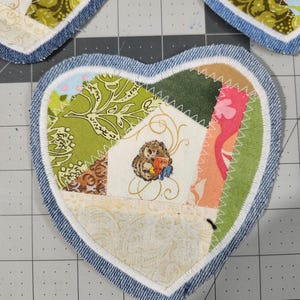 May include: Heart-shaped fabric coasters with a patchwork design. Each coaster features a denim border and a center composed of various patterned fabrics in shades of green, pink, and cream. One coaster has an embroidered owl.