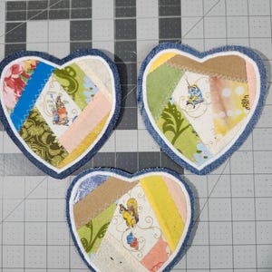 May include: Three heart-shaped fabric coasters with a patchwork design. Each coaster features a denim blue border, a white stitched edge, and a center composed of various patterned fabrics, including floral and animal prints. The coasters are arranged on a gray grid surface.