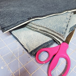 May include: A stack of denim fabric squares, some are light blue and some are dark blue, with a pink pair of scissors on a blue cutting mat.