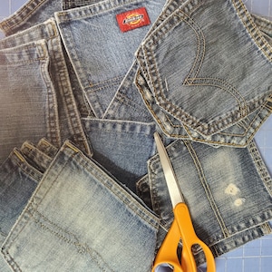 May include: A collection of denim jean pockets in various shades of blue, some with visible stitching and brand labels. An orange-handled pair of scissors rests on top of the pockets, suggesting a craft or DIY project.