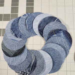 May include: A circular wreath composed of denim circles in varying shades of blue. The denim circles are arranged in a layered pattern, creating a textured and dimensional effect. The wreath is set against a grid background.