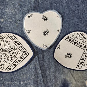 May include: Three white heart-shaped patches with a black paisley pattern. The patches are sewn onto a blue denim fabric.