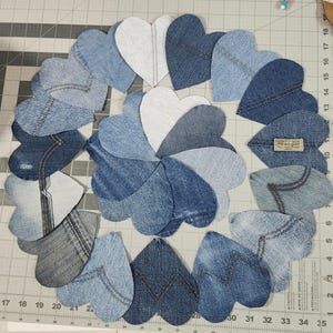 May include: A circular arrangement of denim hearts in various shades of blue and white. The hearts are cut from recycled denim fabric, some with visible stitching and pocket details. A Wrangler label is visible on one heart.