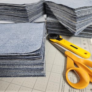 May include: A stack of blue denim fabric squares, cut and ready for a sewing project. A yellow rotary cutter and scissors are on a white cutting mat with black grid lines.