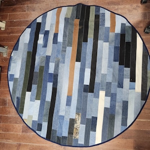 May include: Circular denim rug with a patchwork design. The rug features vertical strips of various shades of blue denim, with accents of black, brown, and white. A dark blue border frames the rug, creating a cohesive look.