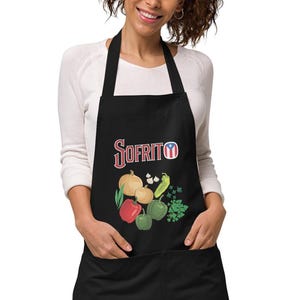Puede incluir: Delantal negro con diseño de Sofrito. El delantal presenta una colorida ilustración de verduras, incluyendo tomates, cebollas y pimientos. La palabra "Sofrito" está escrita en una fuente estilizada.