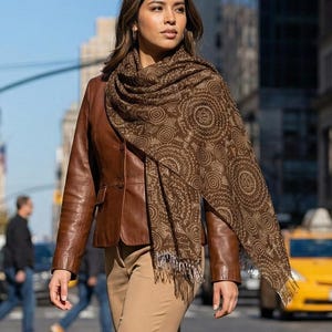 May include: A woman wearing a brown leather jacket, tan pants, and brown heels walks across a city street. She is also wearing a brown scarf with a circular pattern. Yellow taxis and pedestrians are in the background.