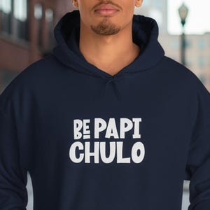 May include: Navy blue hoodie with the white text "BE PAPI CHULO". The hoodie has a drawstring hood and a front kangaroo pocket. The model is wearing the hoodie in an outdoor setting.