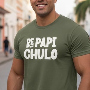 May include: Olive green t-shirt with the text "BE PAPI CHULO" in white block letters. The t-shirt is short-sleeved and appears to be made of a soft material. The person wearing the shirt is smiling and standing on a cobblestone street.