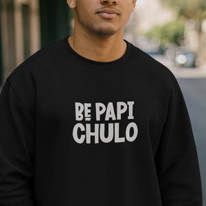 May include: Black sweatshirt with the text "BE PAPI CHULO" in white block letters. The sweatshirt is a crew neck style. The image is taken outdoors with a person wearing the sweatshirt.