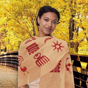 May include: A woman wearing a beige scarf with red symbols and the word "We" printed on it. The scarf is draped around her neck and shoulders. She is standing in front of a wooden bridge with fall foliage in the background.