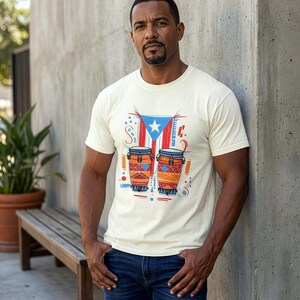 May include: Cream-colored t-shirt featuring a graphic of two conga drums and the Puerto Rican flag. The design includes red, blue, and white elements. The shirt is short-sleeved and worn by a person in blue jeans.