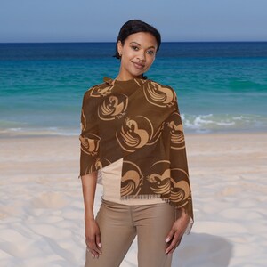 May include: A woman wearing a brown shawl with a tan pattern of stylised birds. The shawl is draped around her shoulders and tied at her neck. She is standing on a sandy beach with the ocean in the background.