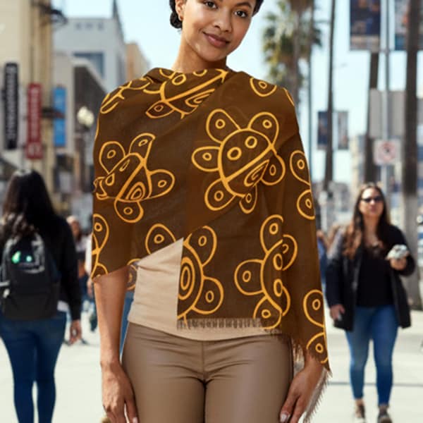 May include: A brown scarf with a repeating pattern of yellow abstract shapes. The scarf is draped over a person's shoulders, showcasing the design. The scarf has a fringed edge.