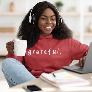 May include: A person wearing a red hoodie with the word "grateful" in white script. They are smiling and wearing white headphones, holding a white mug with pink polka dots, and using a laptop. The scene suggests a relaxed, at-home setting.