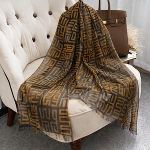 May include: A sheer scarf with a geometric pattern in shades of gold and black is draped over a white armchair. The scarf has a fringed edge. A brown leather handbag sits on a small table in the background.