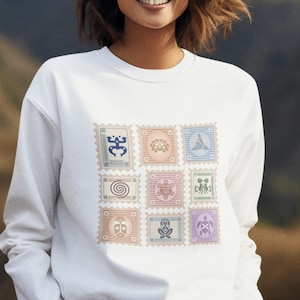 May include: White sweatshirt featuring a grid of nine colorful postage stamp designs. Each stamp showcases a unique, stylized graphic in shades of blue, pink, green, and purple. The sweatshirt has long sleeves and a crew neck.