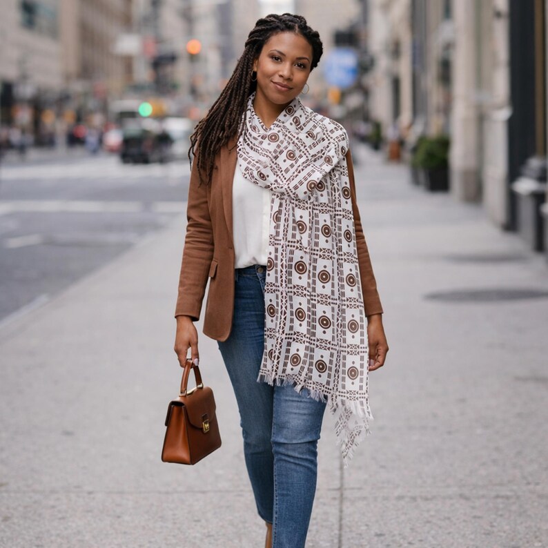 Peut inclure: Une femme porte un blazer marron, un jean bleu et une &eacute;charpe &agrave; motifs. Elle tient un sac &agrave; main marron. L'&eacute;charpe a un fond blanc avec un motif g&eacute;om&eacute;trique marron r&eacute;p&eacute;t&eacute;. En arri&egrave;re-plan, une rue de la ville.