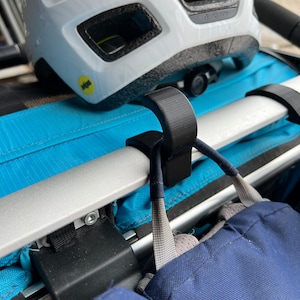 Puede incluir: Un gancho de pl&aacute;stico negro sujeto a una barra plateada. El gancho sostiene una correa azul y gris. Un casco de bicicleta blanco descansa sobre una superficie azul detr&aacute;s del gancho.