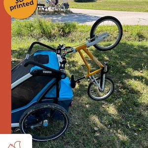 Könnte beinhalten: Ein hellblauer Thule Fahrradanhänger, der über ein 3D-gedrucktes Verbindungsstück an einem gelben 16-Zoll-Woom-Fahrrad befestigt ist. Der Anhänger hat schwarze Netzfenster und einen stabilen Rahmen, während das gelbe Fahrrad hinter dem Anhänger steht.