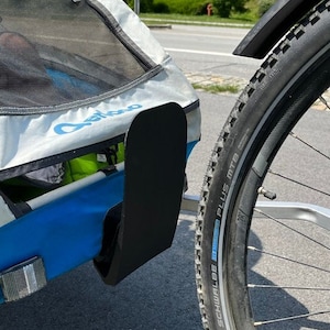 Op de afbeelding: Een blauwe en grijze kinderfietskar met een zwarte spatbord en een reflecterend wit licht. De kar is bevestigd aan een fiets met een zwarte band waarop "Schwalbe Marathon Plus MTB" staat.